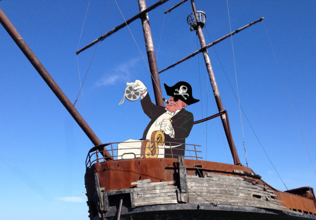 The forward deck of a rigged sailing ship. A ogrish caricatured millionaire stands at a podium sporting a gilded dollar-sign-shaped lever, in place of a ship's wheel. He wears a skull-and-bones pirate hat and eyepatch. He is holding up a fil reel with one white-gloved hand.
Image:
Alan Levine (modified)
https://pxhere.com/en/photo/218986

CC BY 2.0
https://creativecommons.org/licenses/by/2.0/
